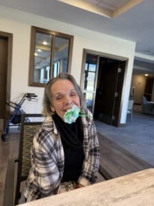 A smiling Meadowview of Davenport memory care resident playfully holds a handmade paper flower in her mouth during an indoor craft activity.